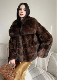 A woman with long dark hair stands indoors, wearing a voluminous, dark brown horizontal-pelt fur jacket with a large shawl collar. She is paired with a shimmering gold satin skirt and holds a tan quilted handbag.