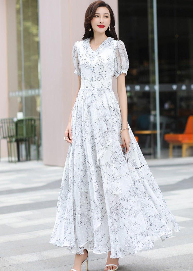 Front-on full-length photo of a chic woman in the Vivian Seven floral print maxi dress, styled with large sunglasses and white sandals. She stands in front of a glass building with outdoor cafe seating.
