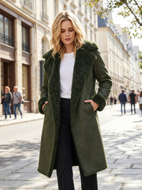 A woman stands on a city street wearing a long olive green shearling-lined coat over a white top and black pants. The background shows blurred buildings and pedestrians.