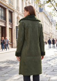 A woman viewed from behind wearing an olive green suede shearling coat with a dark green faux-fur collar and cuffs, standing on a cobblestone street in a city.