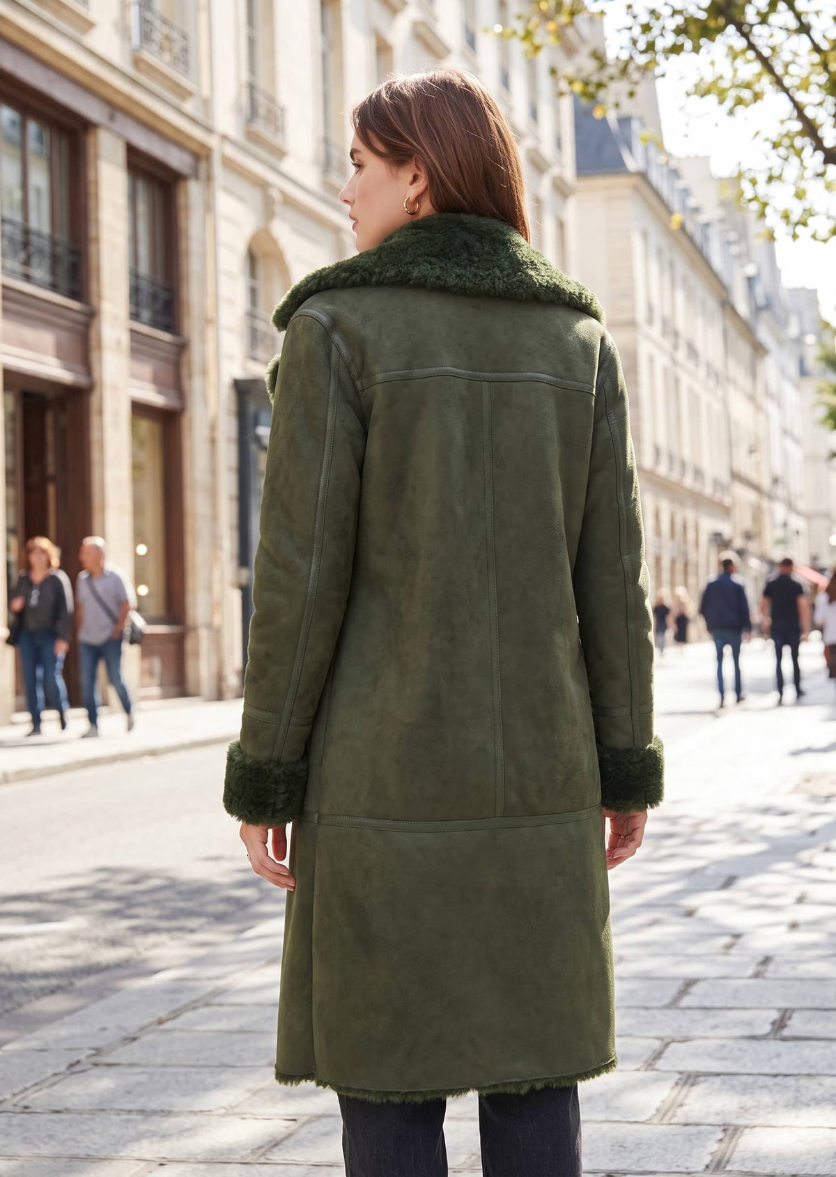 A woman viewed from behind wearing an olive green suede shearling coat with a dark green faux-fur collar and cuffs, standing on a cobblestone street in a city.