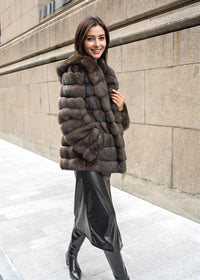 A woman walking on a sidewalk, smiling as she holds the lapel of her horizontal-paneled brown fur coat, paired with a black leather skirt and knee-high boots.