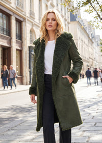 A woman stands on a city street wearing a long olive green shearling-lined coat over a white top and black pants. The background shows blurred buildings and pedestrians.