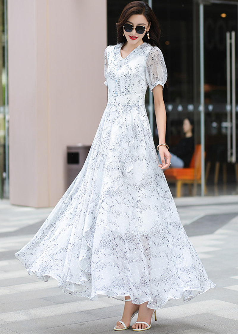 A stylish woman in the full-length Vivian Seven floral chiffon maxi dress with puff sleeves and V-neck. She is posing with confidence in front of a chic, modern building, wearing white sandals and a gold bracelet.