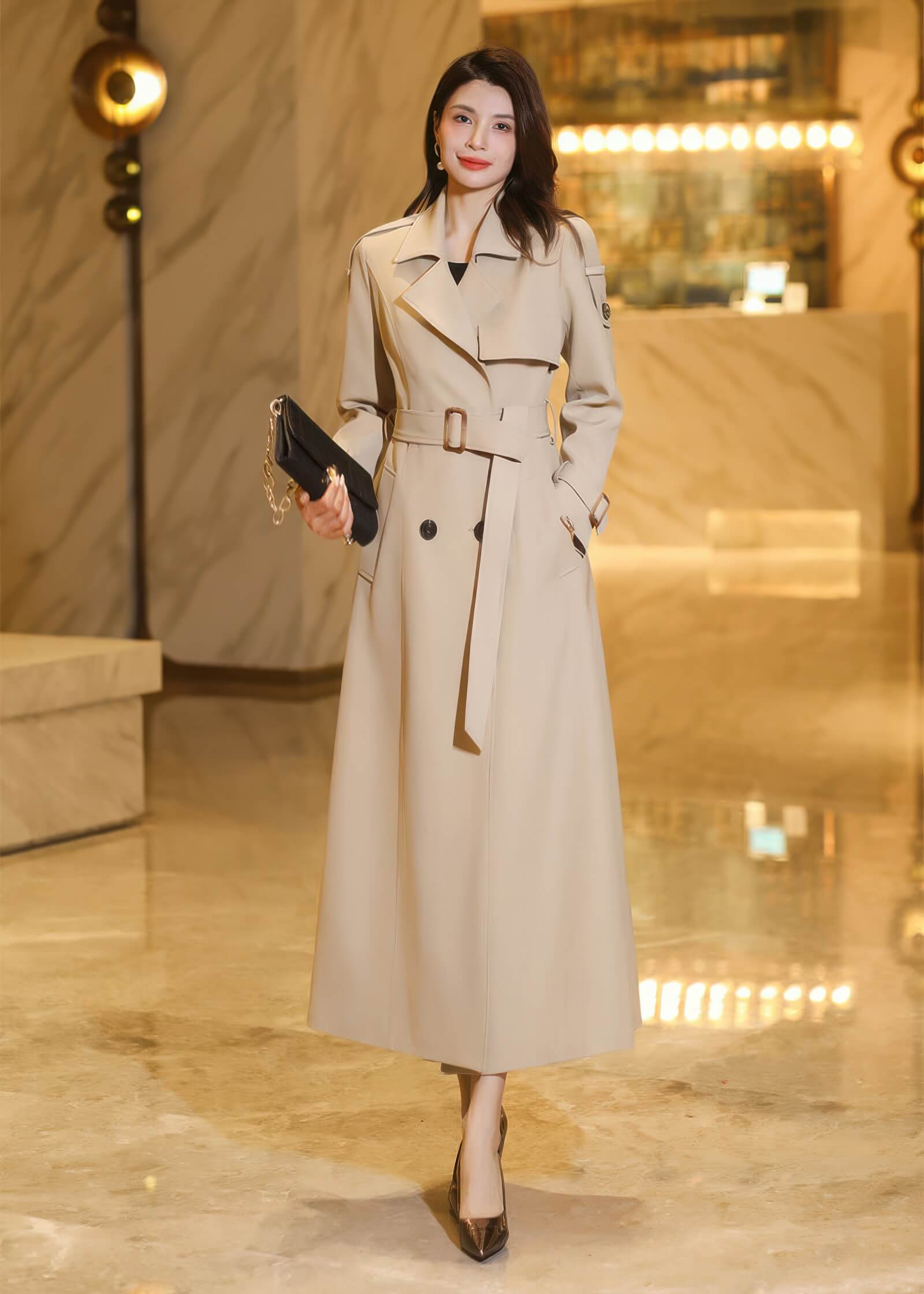 Elegant woman posing in a long beige trench coat holding a black clutch bag in a luxury hotel lobby.