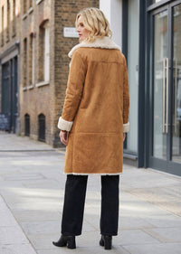 A woman stands on a city sidewalk wearing a camel-brown suede shearling coat, shown from the back. The coat has a cream-colored shearling collar and cuffs.
