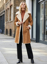 A woman stands on a city sidewalk wearing a tan shearling-lined coat with a large cream collar and cuffs, a rust-brown turtleneck, black wide-leg trousers, and black ankle boots.