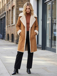 A woman stands on a city sidewalk wearing a mid-length tan shearling coat with a large cream collar and cuffs, a rust-brown turtleneck, black wide-leg trousers, and black ankle boots.
