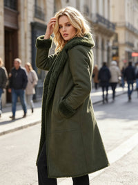 A woman stands on a city street wearing a long olive green coat with a dark green sherpa-lined collar and cuffs, photographed in an outdoor urban setting with blurred pedestrians in the background.