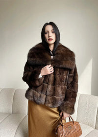 A medium shot of the woman in the brown fur jacket, looking directly at the camera with a neutral expression. She is clutching the front of the jacket with one hand and holding the tan handbag by her side.
