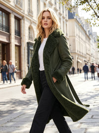 A woman walking on a city street wearing a long olive green shearling-lined coat over a white top and black pants.