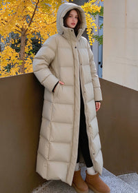 Woman leaning against wall wearing beige 50 inches long hooded duck down puffer coat with hands in pockets