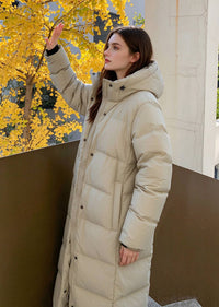 Side profile view of woman raising hand to block sunlight wearing beige 50 inches long hooded puffer coat