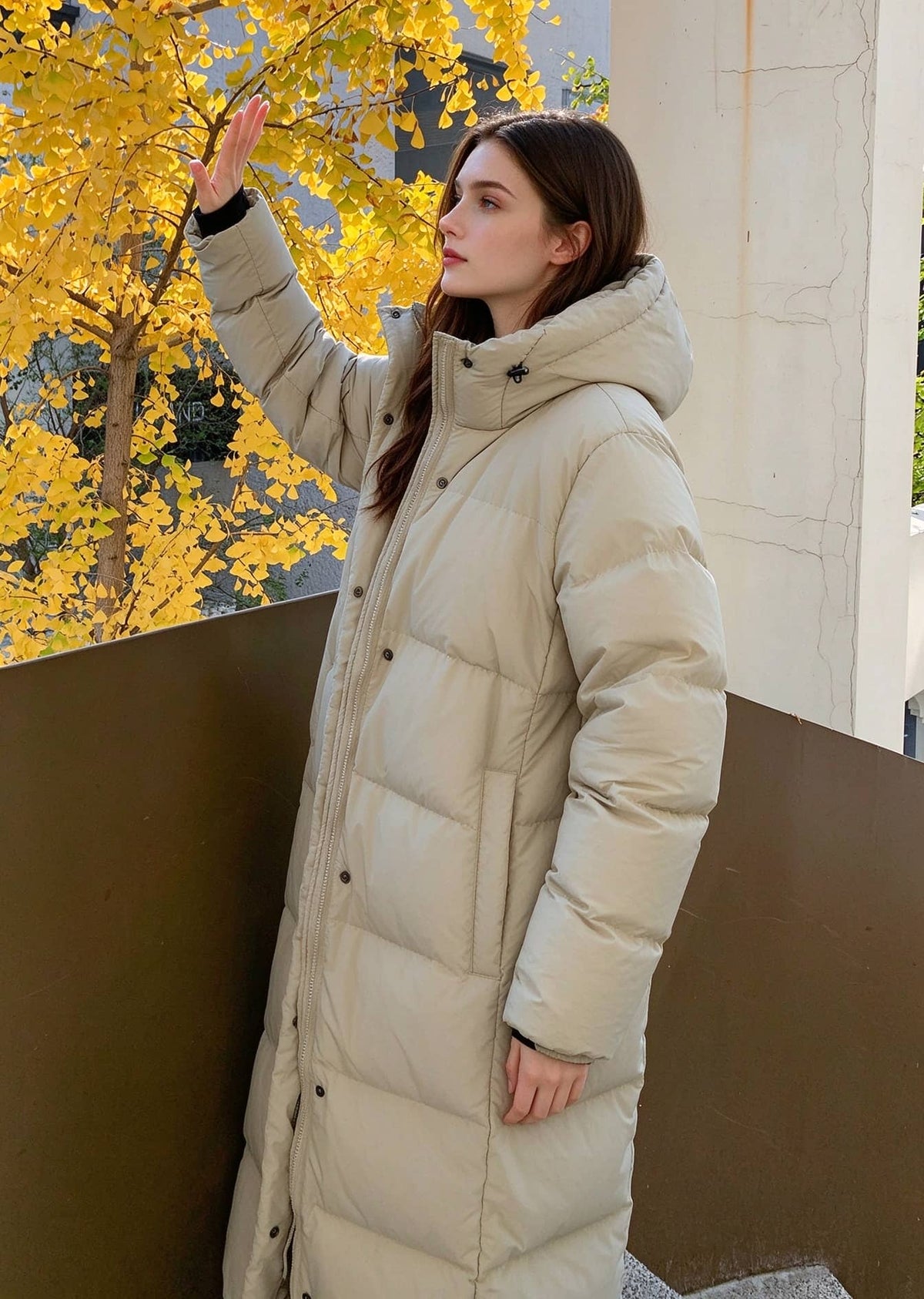 Side profile view of woman raising hand to block sunlight wearing beige 50 inches long hooded puffer coat