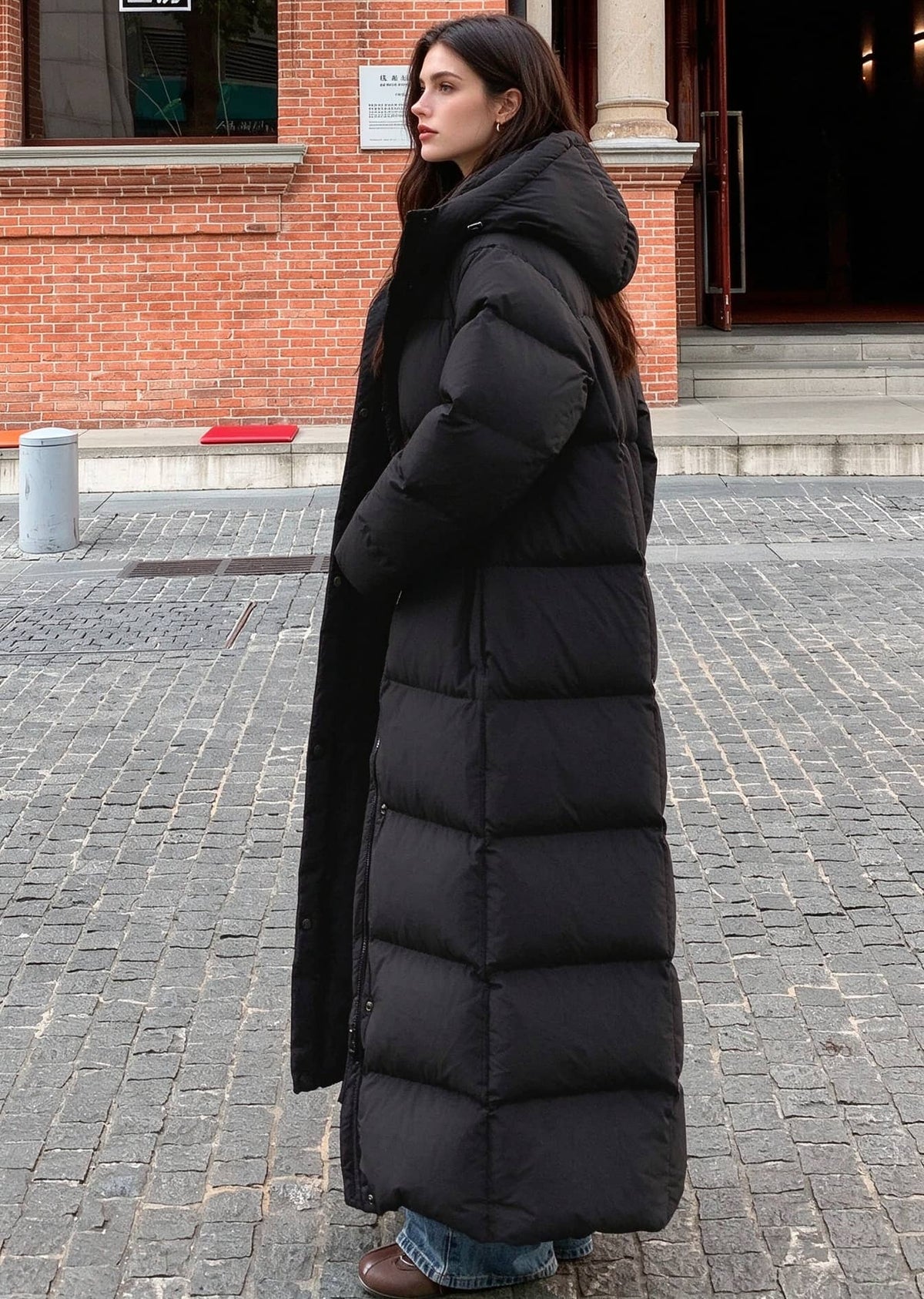 Full length side profile view of woman wearing black 50 inches long hooded duck down puffer coat