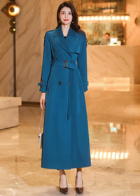 A woman in a buttoned and belted teal long trench coat standing in a luxury interior setting.