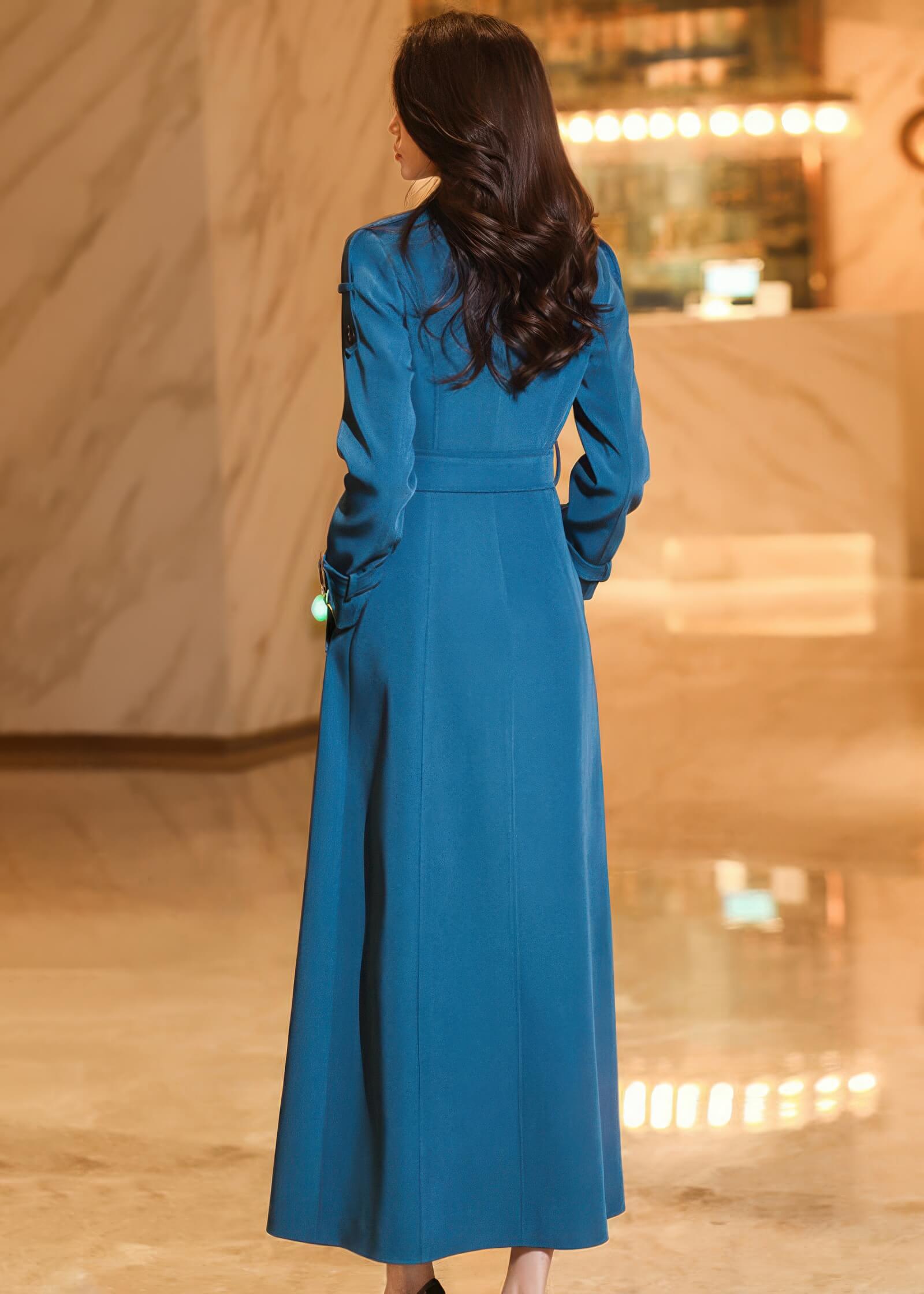 Rear view of a woman wearing a long teal blue trench coat, highlighting the clean silhouette and waist definition.