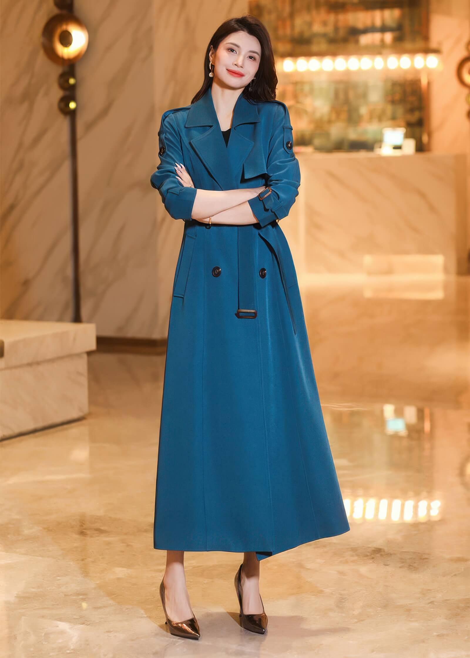 Frontal portrait of a woman in a buttoned-up teal maxi trench coat with arms crossed.