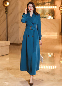 A woman walking in a long teal trench coat styled with a belted waist and a small white handbag.