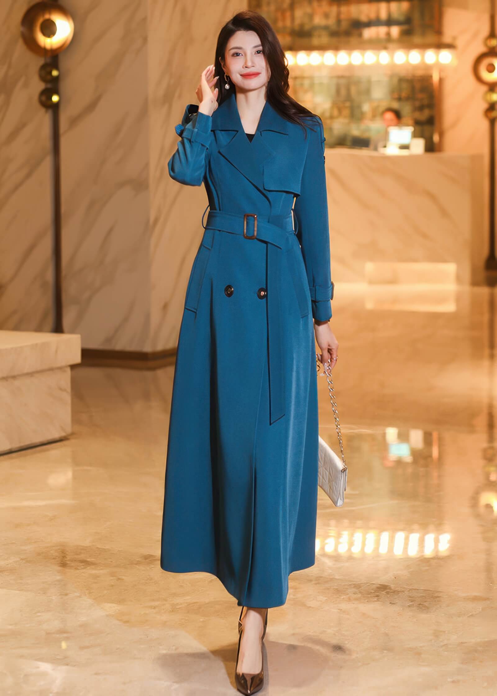 A woman walking in a long teal trench coat styled with a belted waist and a small white handbag.