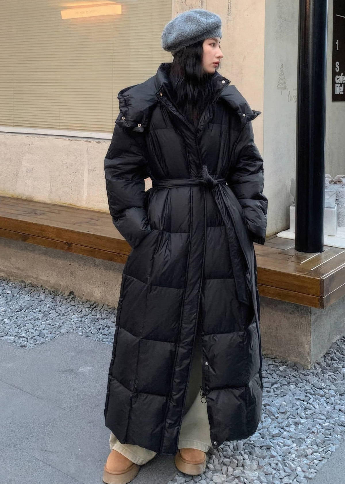 Full-length shot of woman wearing belted black duck down puffer longline coat with neutral winter outfit.