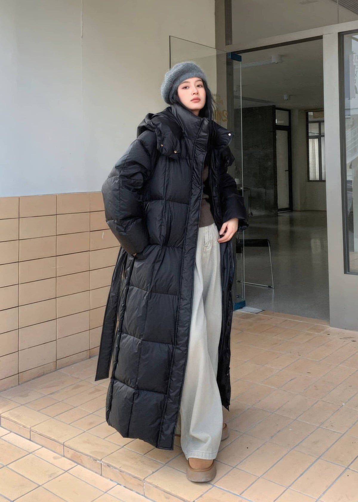 Full-length view of woman wearing open black hooded duck down maxi puffer coat with wide-leg pants.
