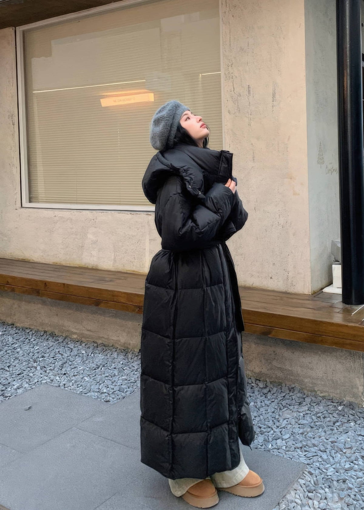Side angle of women's full-length quilted black down puffer coat featuring detachable hood and belt.
