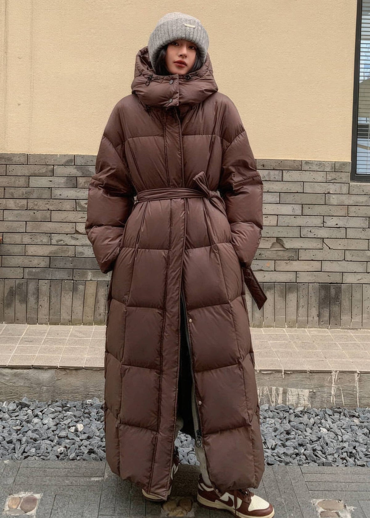 Full-length women's warm brown hooded duck down puffer coat with tied sash belt and ribbed beanie.