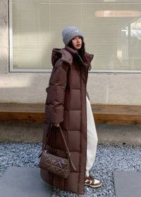 Street style view of woman wearing brown maxi longline puffer coat paired with quilted shoulder bag.