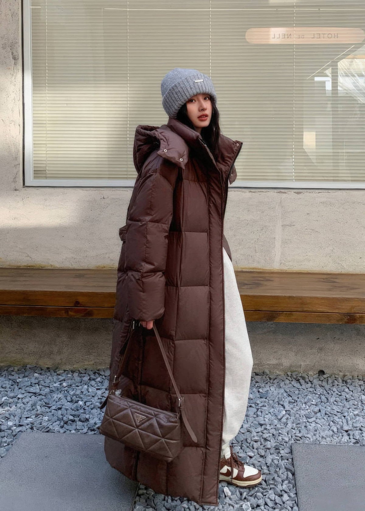 Street style view of woman wearing brown maxi longline puffer coat paired with quilted shoulder bag.