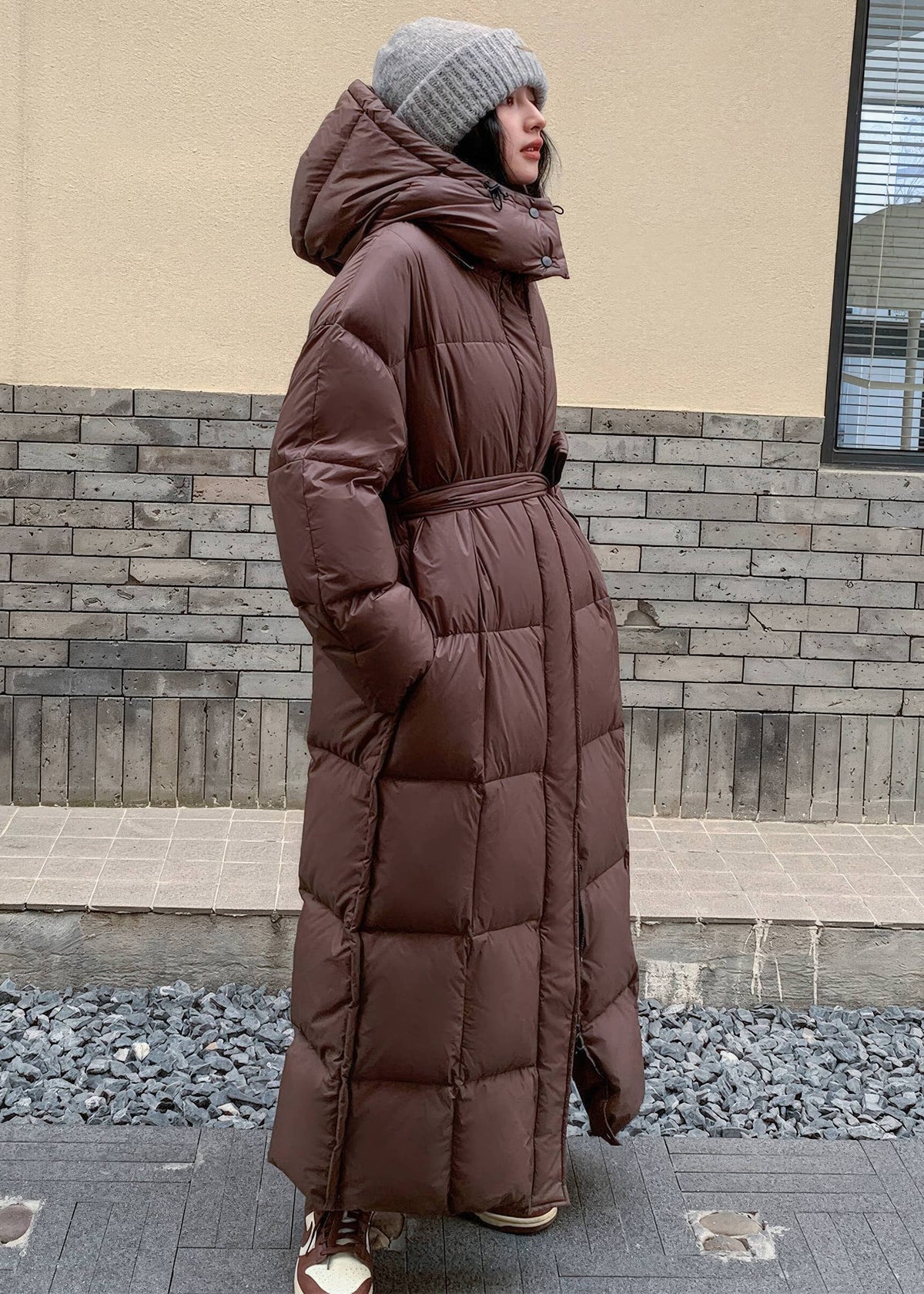 Side profile of women's chocolate brown belted long down jacket with oversized insulated hood.