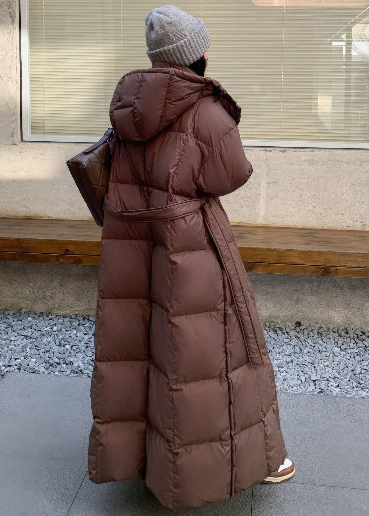 Back view of woman wearing long brown quilted duck down coat showcasing full hood and back belt detail.