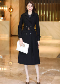 Full-length shot of a woman in a Vivian Seven black double-breasted long trench coat holding a white clutch bag.