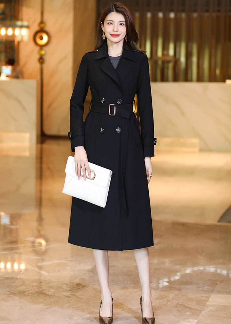 Full-length shot of a woman in a Vivian Seven black double-breasted long trench coat holding a white clutch bag.