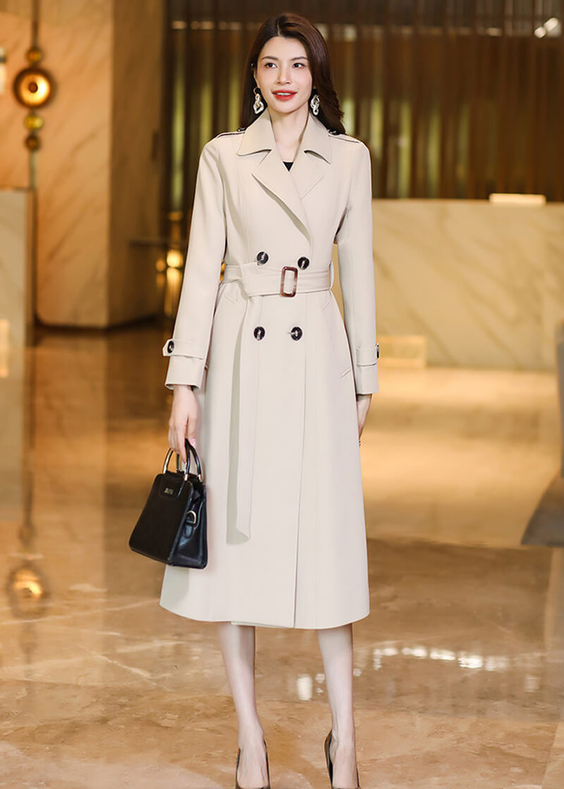 Vivian Seven beige double-breasted long trench coat paired with a black structured handbag and metallic pumps.