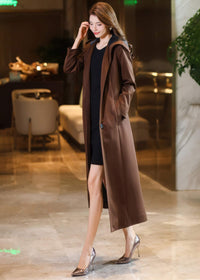 Side profile of a flowing long brown hooded coat with hands in pockets, highlighting the drape and ankle-length cut.
