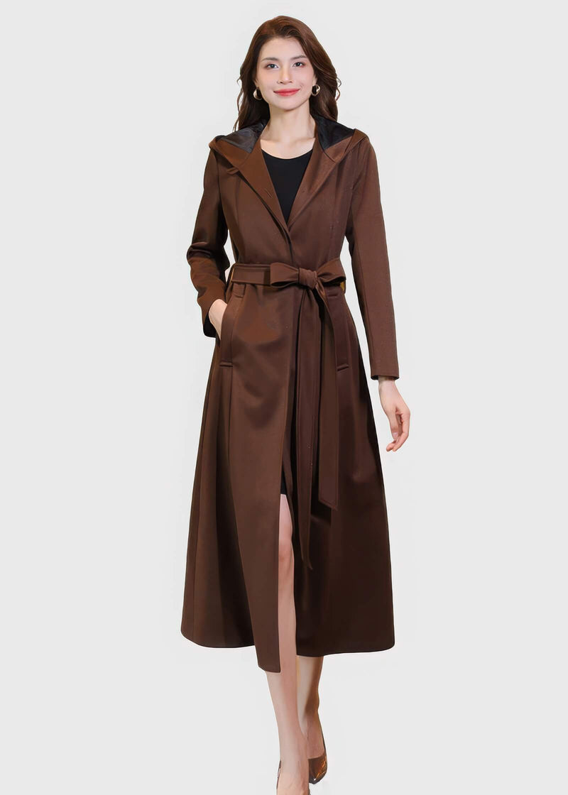 Front view of a women's long brown hooded trench coat worn open with a waist tie, styled over a black dress.
