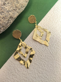 Top-down view of a pair of gold double-diamond drop earrings with a hammered, light-reflecting texture, displayed on a green and white surface.