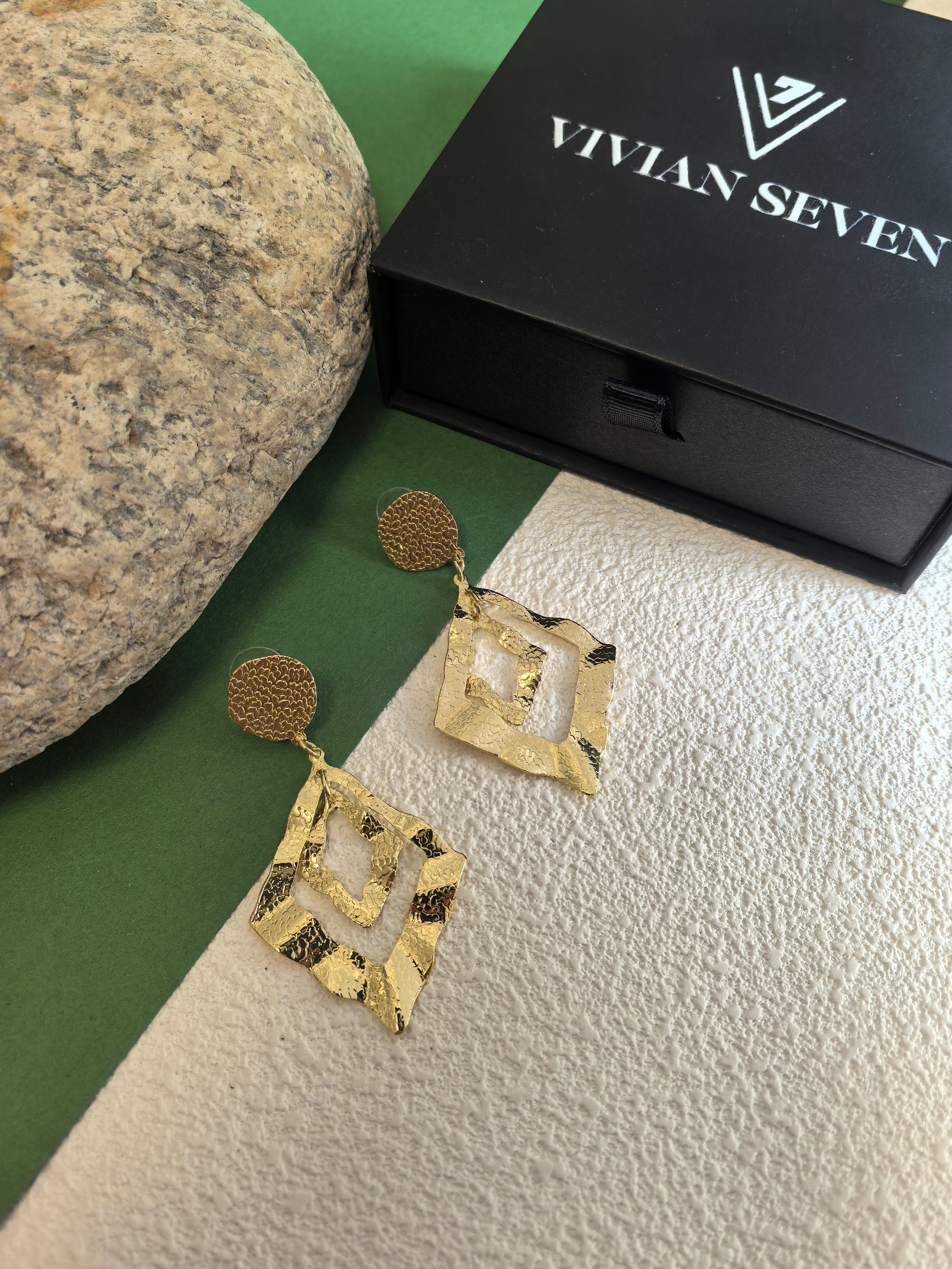 Gold geometric drop earrings displayed on a split green and white textured background next to a black "Vivian Seven" luxury jewelry box.