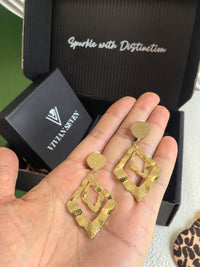 A pair of gold hammered geometric earrings held in a person's open palm to show scale. The background features a black "Vivian Seven" jewelry box with the slogan "Sparkle with Distinction.