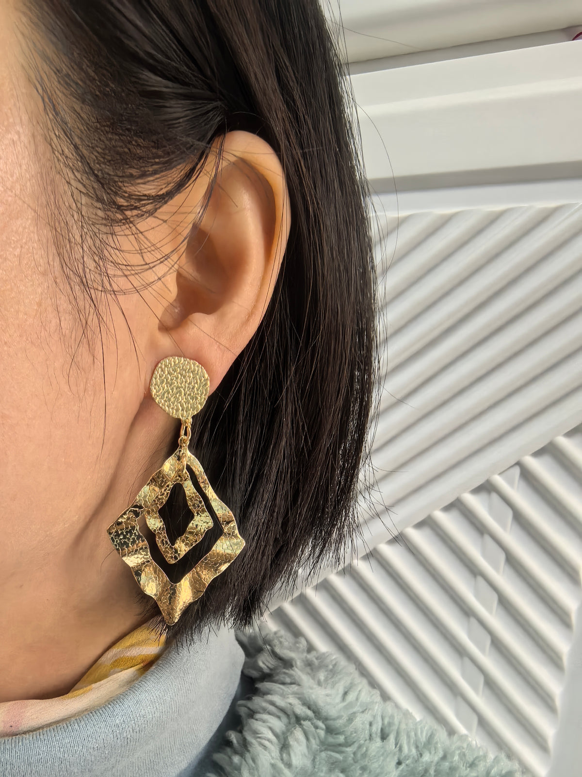 Close-up of a person wearing a large, gold-toned geometric drop earring. The earring features a textured circular stud and a double-layered, hammered metal diamond pendant.