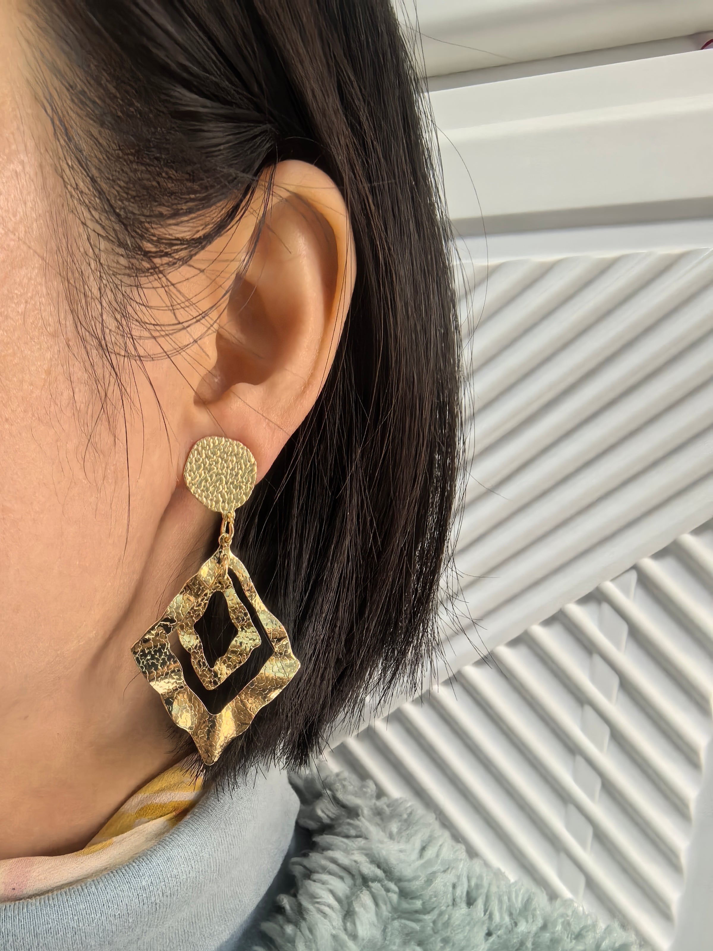 Close-up of a person wearing a large, gold-toned geometric drop earring. The earring features a textured circular stud and a double-layered, hammered metal diamond pendant.