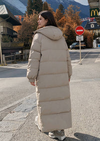 Full length back profile view of woman walking in beige quilted hooded longline polyfill puffer coat