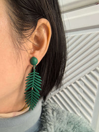 A model wearing one of the dark green, fern-leaf dangle earrings. The earring features a small circular stud and a long, leaf-shaped drop.