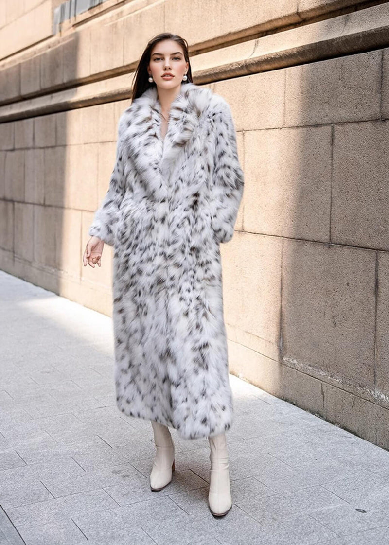 A full-length shot of a woman walking on a sidewalk, wearing a long white faux fur coat with dark spots and pointed-toe cream-colored boots. The background consists of a large stone building.