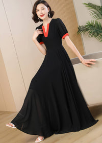 A woman poses wearing a long black A-line maxi dress made of flowing chiffon fabric. The dress features a vibrant red-orange contrast trim around the V-neckline and the cuffs of the short, slightly puffed sleeves. She is leaning gently against a beige sofa, smiling at the camera, and wearing white strappy sandals.