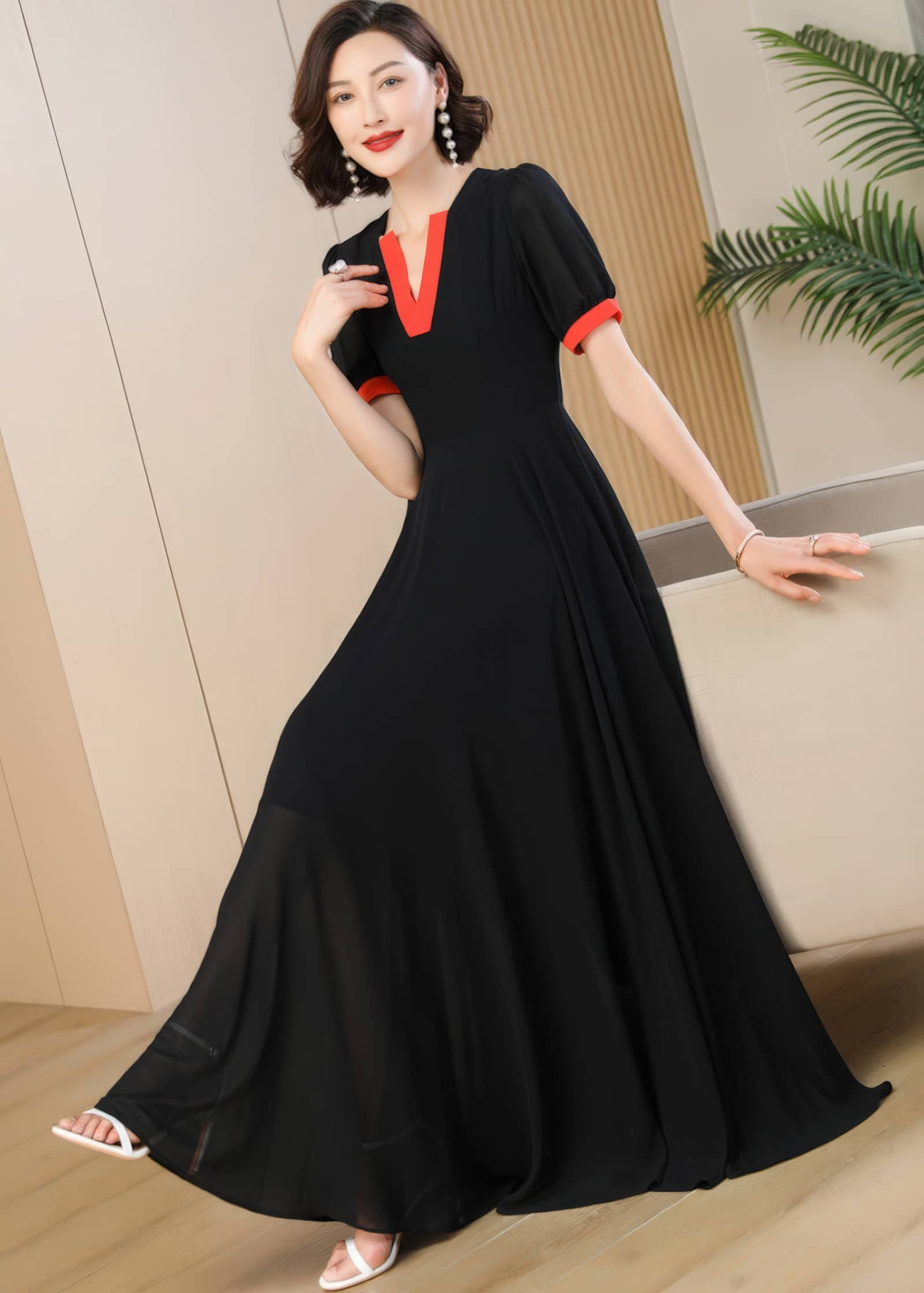 A woman poses wearing a long black A-line maxi dress made of flowing chiffon fabric. The dress features a vibrant red-orange contrast trim around the V-neckline and the cuffs of the short, slightly puffed sleeves. She is leaning gently against a beige sofa, smiling at the camera, and wearing white strappy sandals.