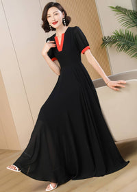 A close-up full-length shot of the woman in the black chiffon maxi dress with orange-red neckline and sleeve accents. She stands with one hand resting on the back of a neutral-toned sofa, showing off the draped silhouette of the skirt and matching pearl drop earrings.