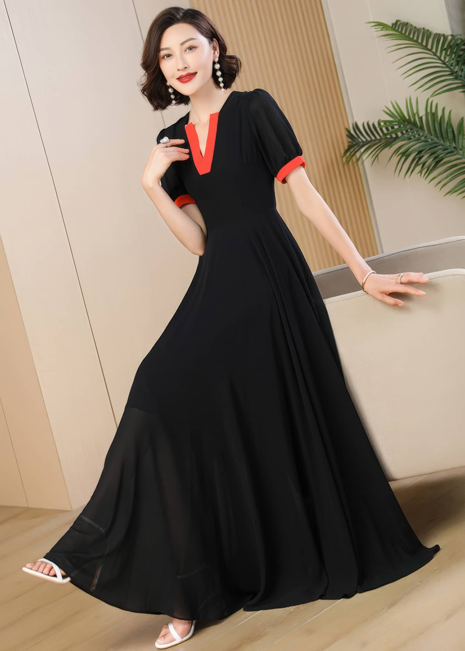 A close-up full-length shot of the woman in the black chiffon maxi dress with orange-red neckline and sleeve accents. She stands with one hand resting on the back of a neutral-toned sofa, showing off the draped silhouette of the skirt and matching pearl drop earrings.