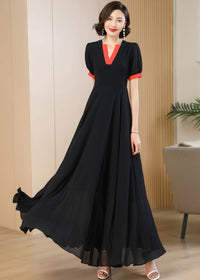 A front-facing view of the woman walking forward in the black maxi dress. The lightweight chiffon fabric of the A-line skirt billows gracefully around her ankles, highlighting its movement. The bold red trim on the V-neck and short sleeves stands out against the dark fabric.
